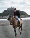 Good Looking Male Horse Rider riding horse on beach in traditional riding clothing with St Michael`s Mount in background Royalty Free Stock Photo