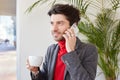 Good looking young glad unshaved brunette male keeping cup of coffee in raised hand and smiling pleasantly, making call while