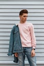 Good-looking young man with hairstyle with stylish blue denim jacket in pink fashionable sweatshirt near white wall in city. Royalty Free Stock Photo