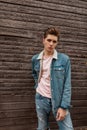 Good-looking young man with hairstyle with stylish blue denim jacket in pink fashionable t-shirt poses near old wooden brown wall Royalty Free Stock Photo