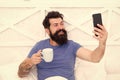 Good morning. Bearded man using mobile technology in bed. Handsome guy talking phone and drinking coffee at home. Happy Royalty Free Stock Photo