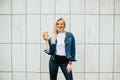 Good morning! Stylish happy young blonde woman wearing stylish jeans jacket, fashion yellow glasses. Cheerful Female holds coffee