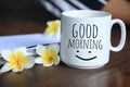 Good morning text greeting on a cup of morning coffee with Bali frangipani flowers, paper work on desk  and an happy smile Royalty Free Stock Photo
