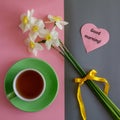 Good morning wishes note in shape heart, tea cup and white yellow daffodils bouquet on pink and grey paper background Royalty Free Stock Photo