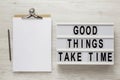 `Good things take time` words on a lightbox, clipboard with blank sheet of paper on a white wooden background, top view. Overhea