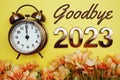 Goodbye 2023 text with alarm clock and flower decoration on yellow background