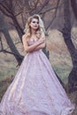 Gorgeous blond lady with luxuriant hairstyle in brocade ball gown standing at the dry leafless tree