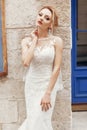 Gorgeous blonde bride in elegant white wedding dress posing near wall outdoors, sexy bride portrait with fancy jewelry and makeup