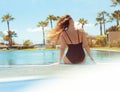 Gorgeous blonde sits near the pool on vacation. A girl with long hair in a single red swimsuit sits on a background of palm trees.