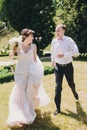 Gorgeous bride in amazing gown and stylish groom running and laughing in sunny park. Beautiful happy wedding couple enjoying time