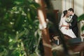 Gorgeous bride with bouquet and stylish groom gently hugging among green leaves in luxury room in hotel. rich wedding couple Royalty Free Stock Photo