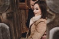 Gorgeous bride in coat and stylish groom gently hugging at old wooden house in winter forest. happy wedding couple embracing on Royalty Free Stock Photo