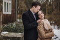 Gorgeous bride in coat and stylish groom posing at wooden house in winter forest. happy wedding couple gently hugging in snowy Royalty Free Stock Photo