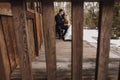 Gorgeous bride in coat and stylish groom sitting at old wooden house in winter forest. happy wedding couple gently hugging on Royalty Free Stock Photo