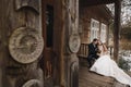 Gorgeous bride in coat and stylish groom sitting at old wooden house in winter forest. happy wedding couple gently hugging on Royalty Free Stock Photo