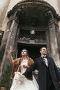 Gorgeous bride in coat and stylish groom throwing candy near church door after wedding ceremony.  happy newlywed couple smiling Royalty Free Stock Photo