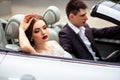 Gorgeous bride with fashion makeup and hairstyle in a luxury wedding dress with handsome groom near white cabriolet car Royalty Free Stock Photo