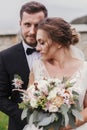 Gorgeous bride with modern bouquet and stylish groom gently hugging and smiling outdoors. Sensual wedding couple embracing. Roman