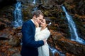 Gorgeous bride softly touches face of her groom. Waterfall on background. Autumn in mountains Royalty Free Stock Photo