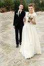 Gorgeous bride and stylish groom holding hands and looking at sandy beach lake, true emotions, luxury wedding Royalty Free Stock Photo