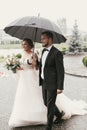 Gorgeous bride and stylish groom walking under umbrella in rainy Royalty Free Stock Photo