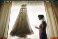 Gorgeous brunette bride with veil looking at vintage white wedding dress hanging on the window, morning wedding preparation, Royalty Free Stock Photo
