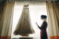 Gorgeous brunette bride with veil looking at vintage white wedding dress hanging on the window, morning wedding preparation, Royalty Free Stock Photo