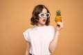 Gorgeous brunette girl in white sunglasses holds pineapple in her hand and looking at it isolated over orange background