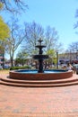 A gorgeous circular red brick water fountain surrounded by people, black metal benches, lush green trees, buildings