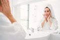 Gorgeous confident adult woman wearing a towel on her head sitting and checking her jaw in front of the mirror. Royalty Free Stock Photo