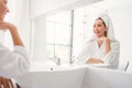 Gorgeous confident middle-aged adult woman wearing a towel on her head sitting and smiling in front of the mirror. Royalty Free Stock Photo