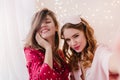 Gorgeous curly woman in sleepmask making selfie in morning with sister. Amazing brunette girl in red pajamas smiling