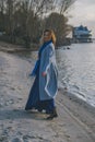 Gorgeous european woman in warm coat and dress on a walk in park near river. Windy weather. Her clothes fly in the wind. Sad,