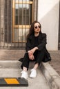 Gorgeous fashion pretty young woman in sunglasses in fashionable black casual wear rests near vintage building on street in city. Royalty Free Stock Photo