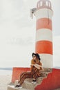 Gorgeous fashionable woman posing while sitting on a lighthouse. Summer photo of a girl in a yellow-orange suit, black hat and