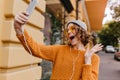 Gorgeous female model in gray hat fooling around on the street while making selfie. Fashionable girl in yellow sweater Royalty Free Stock Photo