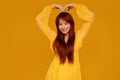 Gorgeous ginger woman in yellow elegant dress smiling to the camera, showing heart symbol. Female bright look. Colorful background