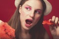 Gorgeous girl with pink make up, wearing jeans, hat and top, posing at red studio background, holding slice watermelon, looking on Royalty Free Stock Photo