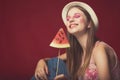 Gorgeous girl with pink make up, wearing jeans, hat and top, posing at red studio background, holding slice watermelon on stick, Royalty Free Stock Photo