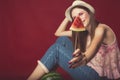 Gorgeous girl with pink make up, wearing jeans, hat and top, posing at red studio background, holding slice watermelon on stick, Royalty Free Stock Photo