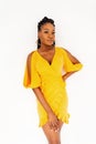 Gorgeous modern young African woman with cool dreadlocks in fashionable yellow dress stands near white wall in studio. Attractive