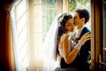 The gorgeous newlywed couple is tenderly hugging near the old open window. The handsome groom is softly kissing his