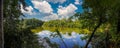 A gorgeous panoramic shot of a still lake surrounded by lush green trees reflecting off the water with blue sky