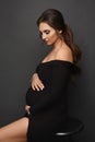 A gorgeous pregnant model girl wearing a black evening dress posing over grey background. Young pregnant woman in