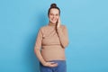 Gorgeous pregnant woman talking phone while posing isolated over blue background, looking at camera smiling, touching her belly Royalty Free Stock Photo