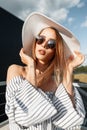 Gorgeous pretty young woman fashion model in beautiful straw hat in elegant striped dress in round trendy sunglasses stands on Royalty Free Stock Photo