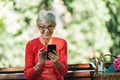 Smiling caucasian senior woman with blonde short hair sitting on bench in park and make call with smart phone Royalty Free Stock Photo