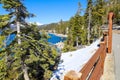 A gorgeous smooth concrete paved hiking trail with a burnt orange wooden fence along the trail with lush green pine trees and snow