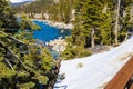 A gorgeous smooth concrete paved hiking trail with a burnt orange wooden fence along the trail with lush green pine trees and snow