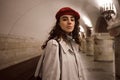 Gorgeous stylish girl in casual trench coat and red beret confidently looking in camera waiting train at metro station Royalty Free Stock Photo